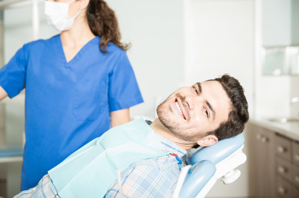A relaxed male patient smiling in a dental chair, with a dental professional in scrubs and a mask in the background, illustrating comfort options like sedation dentistry vs. local anesthesia.